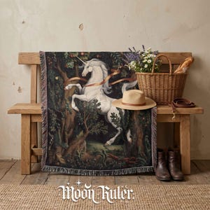 May include: A woven throw blanket featuring a white unicorn in a dark forest setting with green foliage and orange ribbons. The blanket has a fringed edge and is draped over a wooden bench next to a wicker basket, a straw hat, and brown boots.
