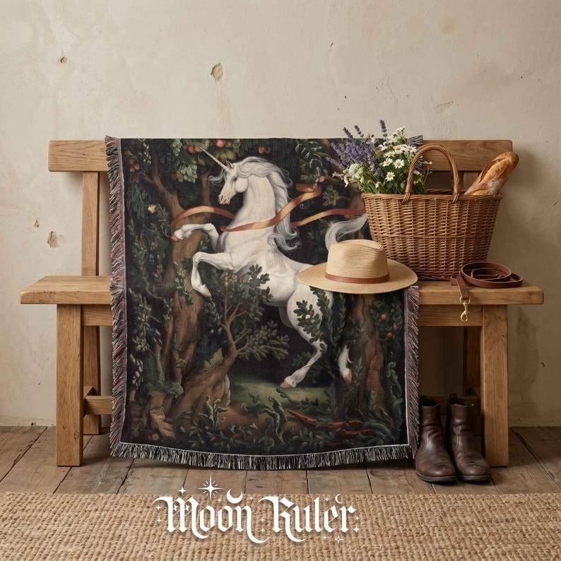 May include: A woven throw blanket featuring a white unicorn in a dark forest setting with green foliage and orange ribbons. The blanket has a fringed edge and is draped over a wooden bench next to a wicker basket, a straw hat, and brown boots.