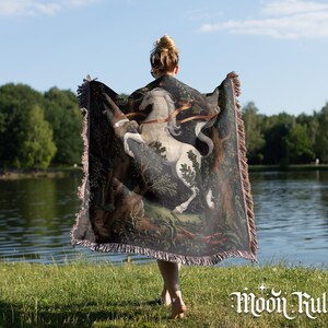 May include: A woven throw blanket featuring a detailed illustration of a white unicorn rearing up in a dark forest setting. The rectangular blanket has fringed edges on all sides and displays a rich color palette of deep greens, browns, and white.
