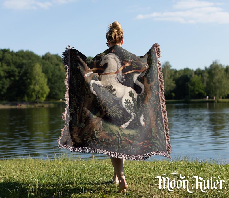 May include: A woven throw blanket featuring a detailed illustration of a white unicorn rearing up in a dark forest setting. The rectangular blanket has fringed edges on all sides and displays a rich color palette of deep greens, browns, and white.