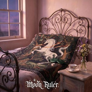 May include: A woven throw blanket featuring a white unicorn in a dark forest setting, draped over a bed with a metal headboard. The text "Moon Ruler" is printed in white at the bottom. A green sleep mask and a teacup are on a side table.
