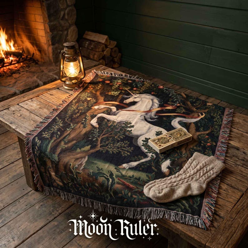 May include: A woven throw blanket featuring a white unicorn in a dark forest setting with trees and foliage. The blanket has a fringed edge and is displayed with a pair of beige cable-knit socks and a small rectangular box on top of it.