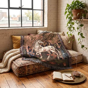 May include: A woven throw blanket featuring a detailed illustration of a white unicorn standing in a dark, lush forest with red flowers and green foliage. The rectangular blanket has a fringed edge and a dark, moody colour palette throughout.