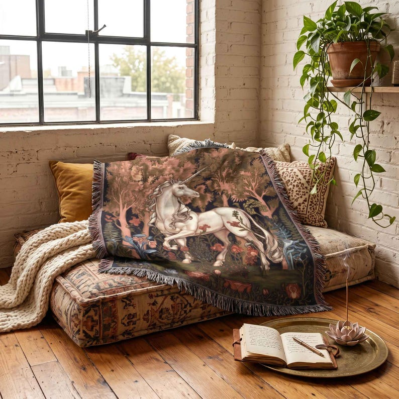 May include: A woven throw blanket featuring a detailed illustration of a white unicorn standing in a dark, lush forest with red flowers and green foliage. The rectangular blanket has a fringed edge and a dark, moody colour palette throughout.
