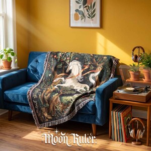 May include: A woven throw blanket featuring a detailed illustration of a white unicorn in a dark forest setting with trees and foliage. The blanket has a fringed edge and is draped over a blue sofa. The text Moon Ruler is visible at the bottom.