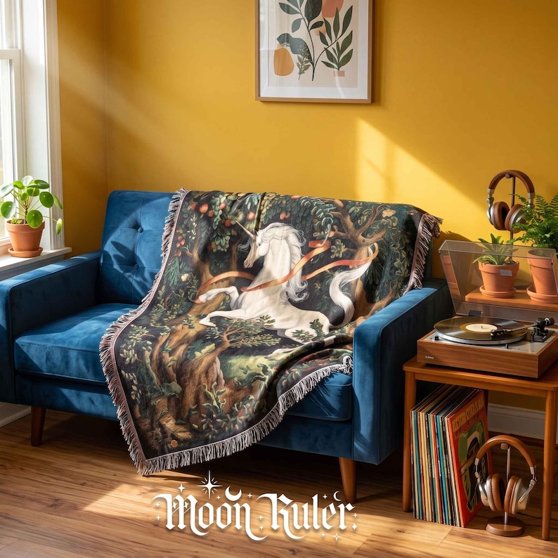 May include: A woven throw blanket featuring a detailed illustration of a white unicorn in a dark forest setting with trees and foliage. The blanket has a fringed edge and is draped over a blue sofa. The text Moon Ruler is visible at the bottom.
