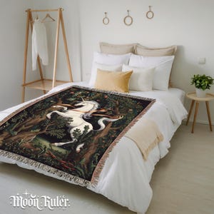 May include: A woven throw blanket featuring a detailed illustration of a white unicorn standing in a dark, lush forest with trees and foliage. The blanket has a fringed edge and is draped over a bed with white bedding and several pillows.
