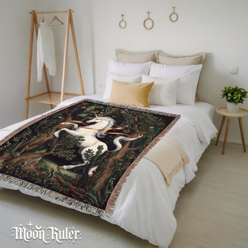 May include: A woven throw blanket featuring a detailed illustration of a white unicorn standing in a dark, lush forest with trees and foliage. The blanket has a fringed edge and is draped over a bed with white bedding and several pillows.
