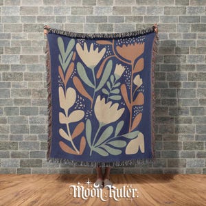 Puede incluir: Una manta azul marino con un diseño floral en beige, verde y óxido. La manta tiene un borde con flecos y el texto "Moon Ruler" en la parte inferior.