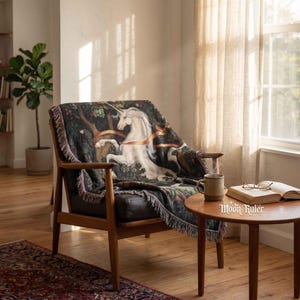 May include: A woven throw blanket featuring a white unicorn illustration set against a dark forest background with orange ribbons, finished with a fringed edge. The blanket is draped over a wooden armchair with a dark seat cushion.