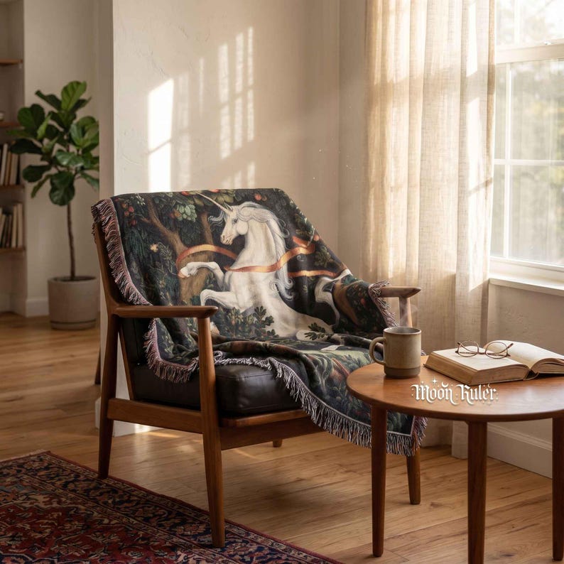 May include: A woven throw blanket featuring a white unicorn illustration set against a dark forest background with orange ribbons, finished with a fringed edge. The blanket is draped over a wooden armchair with a dark seat cushion.