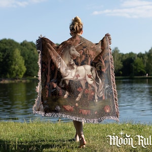May include: A woven throw blanket featuring a detailed illustration of a white unicorn standing in a forest with flowers and trees. The blanket has a fringed edge and displays the text Moon Ruler in white lettering at the bottom right corner.