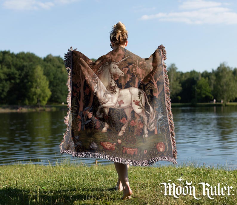 May include: A woven throw blanket featuring a detailed illustration of a white unicorn standing in a forest with flowers and trees. The blanket has a fringed edge and displays the text Moon Ruler in white lettering at the bottom right corner.