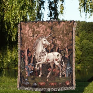 May include: A rectangular woven throw blanket featuring a detailed illustration of a white unicorn standing in a dark forest with reddish-brown trees and various flowers. The blanket has a fringed edge and the text Moon Ruler at the bottom.