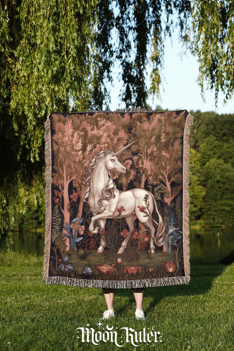 May include: A rectangular woven throw blanket featuring a detailed illustration of a white unicorn standing in a dark forest with reddish-brown trees and various flowers. The blanket has a fringed edge and the text Moon Ruler at the bottom.