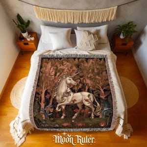 May include: A woven throw blanket featuring a detailed illustration of a white unicorn standing in a dense forest with reddish-brown trees and foliage. The blanket has a fringed edge and is draped over a bed with white pillows and a cream-coloured cover.