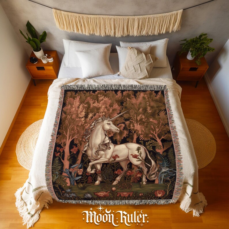 May include: A woven throw blanket featuring a detailed illustration of a white unicorn standing in a dense forest with reddish-brown trees and foliage. The blanket has a fringed edge and is draped over a bed with white pillows and a cream-coloured cover.