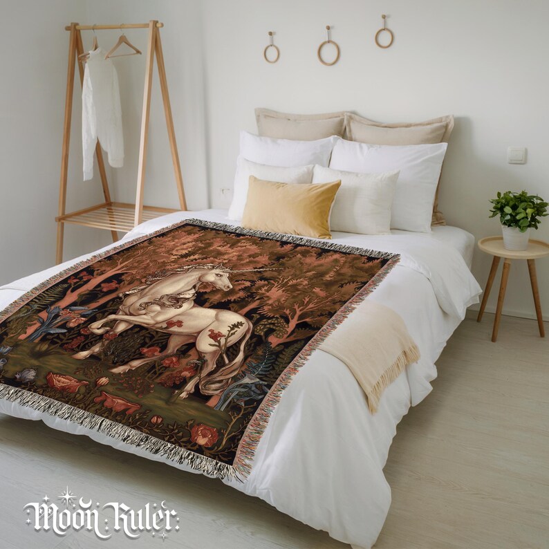 May include: A woven throw blanket featuring a detailed depiction of a white unicorn standing in a dense forest with dark green foliage and red flowers. The blanket has a fringed edge and is draped over a white bedspread in a bright bedroom.
