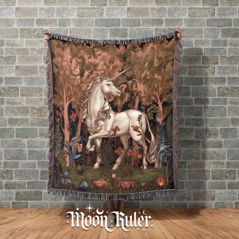May include: A woven throw blanket featuring a white unicorn standing in a forest with pink and brown trees, green foliage, and red flowers. The rectangular blanket has a fringed edge and a detailed tapestry-style design with a dark background.