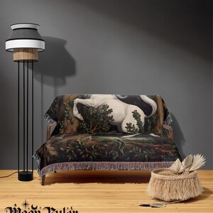 May include: A woven throw blanket featuring a detailed tapestry-style illustration of a white unicorn in a dark forest setting, finished with decorative fringe along the edges. The blanket is draped over a wooden sofa next to a tall floor lamp.