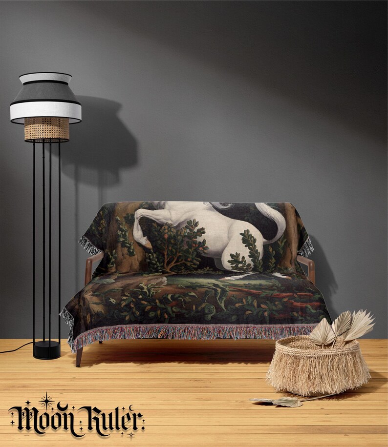 May include: A woven throw blanket featuring a detailed tapestry-style illustration of a white unicorn in a dark forest setting, finished with decorative fringe along the edges. The blanket is draped over a wooden sofa next to a tall floor lamp.
