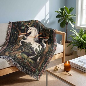 May include: A woven throw blanket featuring a detailed illustration of a white unicorn standing in a dark forest with trees and foliage. The blanket has a decorative fringed edge and displays a flowing orange ribbon draped across the unicorn.