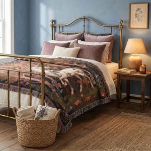 May include: A brass metal bed frame with a woven throw blanket featuring a white unicorn in a forest scene. The bed is dressed with mauve and cream pillows. A round woven basket containing folded cream linens sits in the foreground.