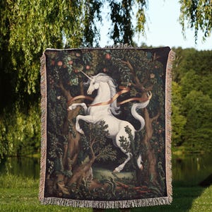 May include: A rectangular woven throw blanket featuring a white unicorn standing in a dark forest with trees, fruit, and a flowing ribbon. The blanket has a fringed edge on all sides and displays the text Moon Ruler at the bottom center.