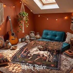 May include: A woven throw blanket featuring a white unicorn in a forest setting with red flowers and green foliage. The blanket has a fringed edge and displays the text Moon Ruler in a white decorative font at the bottom centre of the design.