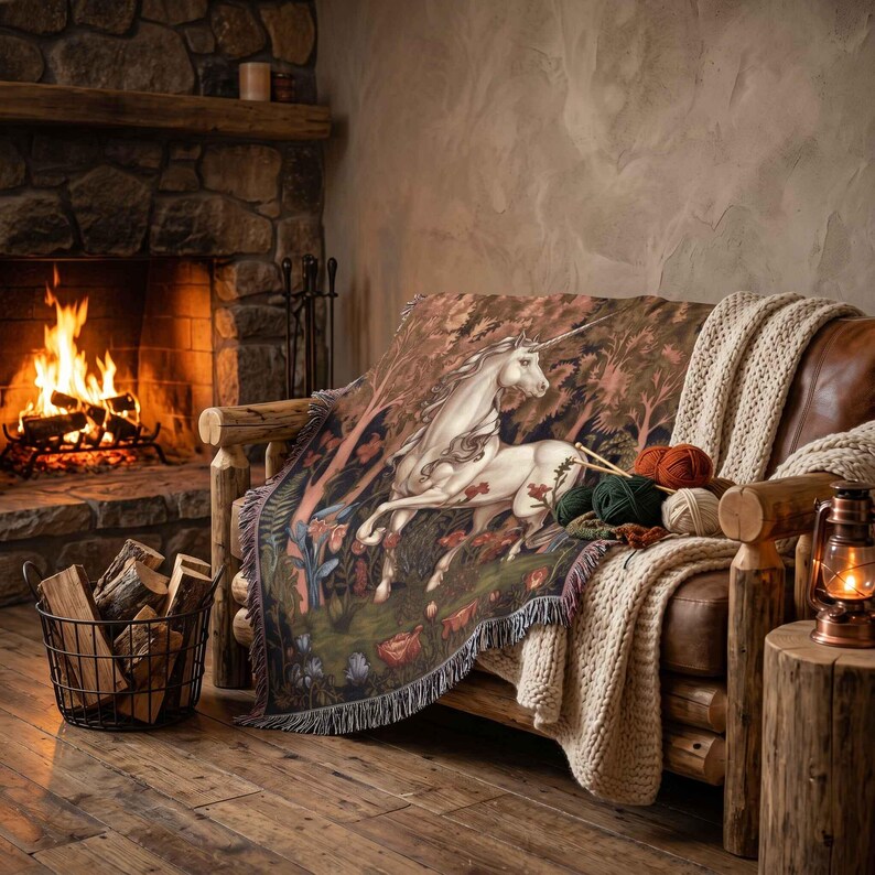 May include: A woven throw blanket featuring a detailed illustration of a white unicorn in a forest setting with flowers and trees. The blanket has a fringed edge and is draped over a brown leather sofa alongside several balls of yarn.