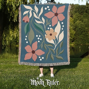 May include: A teal throw blanket with a floral design featuring pink, tan, and cream-coloured flowers and leaves. The blanket has a fringed edge and the words "Moon Ruler" in a stylized font at the bottom.