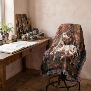May include: A woven throw blanket featuring a detailed illustration of a white unicorn standing in a lush forest with red flowers and green foliage. The blanket has a fringed edge and is draped over a dark metal chair in a studio setting.