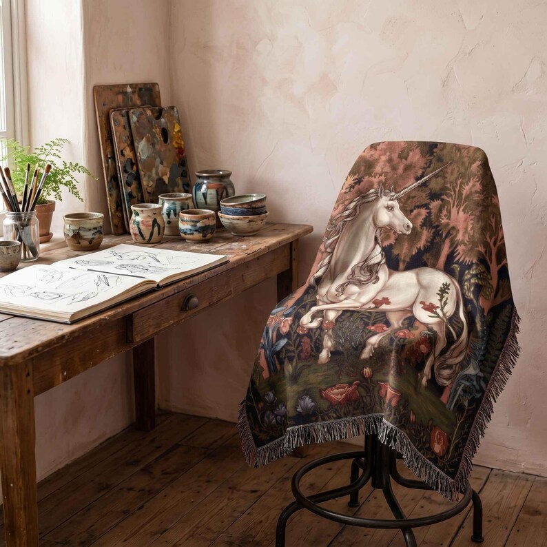 May include: A woven throw blanket featuring a detailed illustration of a white unicorn standing in a lush forest with red flowers and green foliage. The blanket has a fringed edge and is draped over a dark metal chair in a studio setting.