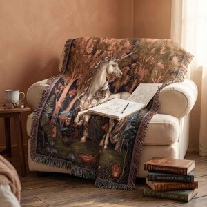 May include: A woven throw blanket featuring a detailed illustration of a white unicorn in a forest setting with trees and flowers. The blanket has a fringed edge and is draped over a cream-coloured armchair. An open sketchbook rests on top.