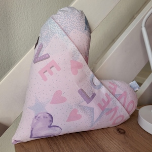 Peut inclure: Un coussin en tissu en forme de cœur avec un fond rose clair orné d'un motif répétitif de cœurs, d'étoiles et du mot LOVE imprimé en grandes lettres majuscules violettes et roses. Le textile présente une texture pointillée sur tout le motif.