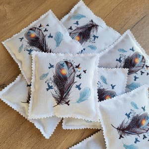 Può includere: Una collezione di piccoli sacchetti quadrati di tessuto bianco con un design decorativo di piume e uccelli. La piuma è principalmente marrone scuro e nera con un centro giallo e arancione. Uccelli e piume blu sono sparsi intorno al disegno principale. I bordi dei sacchetti hanno un bordo smerlato.