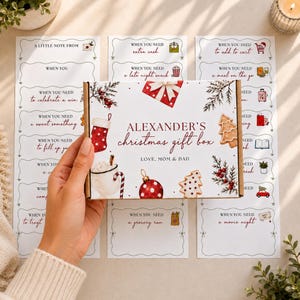 May include: A rectangular white cardboard gift box labeled Alexander's Christmas gift box, Love, Mom & Dad, decorated with illustrations of a red stocking, a mug, a red ornament, and cookies, surrounded by several white cards with holiday text.