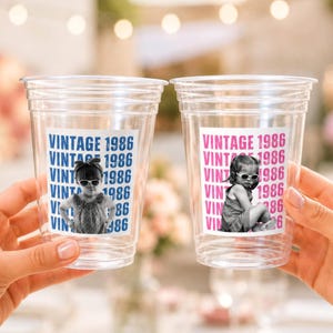 May include: Two clear plastic disposable cups featuring a printed label with the text VINTAGE 1986 repeated in the background. The left cup displays a black and white photo of a child wearing sunglasses, while the right cup shows a similar child.
