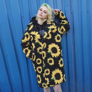 Puede incluir: Abrigo largo negro con estampado de girasoles amarillos. El abrigo tiene capucha y cierre de cremallera. La persona lleva una sudadera con capucha negra. El fondo es una pared azul lisa.