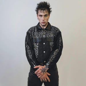 Könnte beinhalten: Schwarze Jacke mit silbernen Verzierungen und Lederakzenten. Die Jacke hat eine Knopfleiste, zwei Brusttaschen und einen Kragen. Die Ärmel und die obere Brust sind mit silbernen Nieten verziert. Das Model trägt eine schwarze Lederhose.