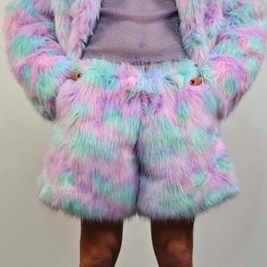 以下が含まれることがあります： 鮮やかな色のフェイクファーのジャケットとショートパンツのセット。 服はパステルカラーのピンク、紫、ターコイズのミックスです。 この衣装は柔らかくふわふわした質感で、独特で人目を引く外観をしています。