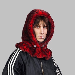 Può includere: Una persona indossa un collo di pelliccia sintetica rosso con cappuccio, una giacca nera e strisce bianche. Il collo ha una chiusura a bottone.