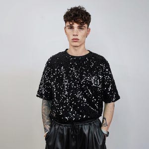 May include: Black sequin t-shirt with short sleeves. The shirt is covered in shiny, reflective sequins. The model is wearing black leather pants with a drawstring waist. The background is a plain, light gray.