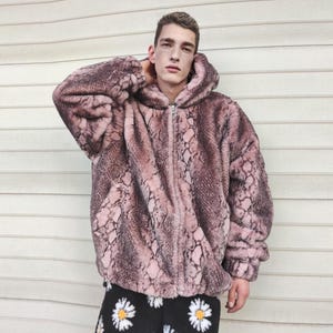May include: A hooded zip-up jacket with a snakeskin pattern in shades of pink and brown. The jacket is made of a soft, plush material. The model is wearing black pants with white daisy patterns.
