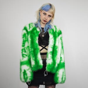 May include: A bright green and white faux fur jacket with a tie-dye effect. The jacket has a fluffy texture. The model is wearing a black top and skirt, with fishnet stockings. The background is plain white.