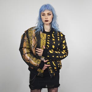 Może przedstawiać: Czarna kurtka bomberka o unikalnym designie. Jedna strona ma wzór skóry węża w odcieniach żółci, brązu i czerni, a druga strona ma czarne tło z żółtymi abstrakcyjnymi kształtami. Kurtka ma czarny kołnierz i mankiety.