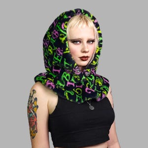 May include: A black hooded neck warmer with a green and pink print of letters and symbols. The model is wearing the neck warmer and has tattoos on their arms.