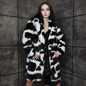 Può includere: Un lungo cappotto in finta pelliccia con un motivo a stampa di mucca in bianco e nero. Il cappotto è indossato sopra un vestito nero e abbinato a stivali neri. Il design del cappotto presenta forme nere irregolari su sfondo bianco.