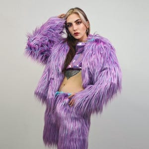 Könnte beinhalten: Ein auffälliges Set aus lila Kunstfelljacke und Hose. Die Jacke hat lange Ärmel und eine zottelige Textur. Das Outfit wird durch ein paillettenbesetztes Crop-Top ergänzt. Dieses Ensemble ist ein mutiges Mode-Statement.