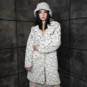 May include: A white faux fur coat and matching bucket hat with a black pattern. The long coat has a notched collar and a button closure. The hat has a rounded crown and a wide brim. The outfit is worn over a white dress.